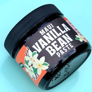 May include: A small, round, dark glass jar with a black screw-top lid containing Maui Vanilla Bean Paste. The label is black and orange with white text and illustrations of white vanilla flowers with green leaves on the front of the container.