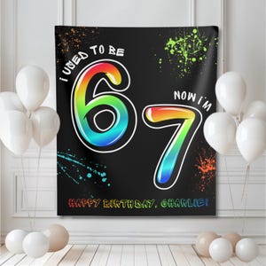 Pode incluir: Uma faixa de aniversário preta com o texto "I USED TO BE 67 NOW I'M 7" em uma fonte gradiente colorida. A faixa também inclui o texto "HAPPY BIRTHDAY, CHARLIE!" e respingos de tinta coloridos. Balões brancos em primeiro plano.