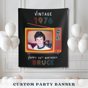 Puede incluir: Bandera negra con un gráfico de TV vintage con la foto de una persona. La bandera dice "VINTAGE 1976" y "Happy 50th Birthday, Bruce". Globos blancos en el fondo. La parte inferior de la bandera dice "CUSTOM PARTY BANNER."