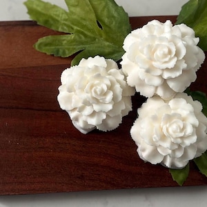 May include: Three white carnation-shaped soap bars on a wooden surface with green leaves.