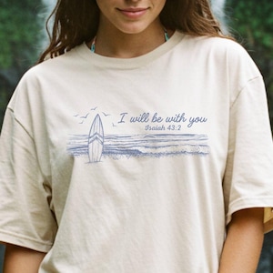 May include: Beige t-shirt with a blue and white graphic of a surfboard and the text "I will be with you" and "Isaiah 43:2". The shirt is worn in front of a waterfall.