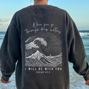 May include: Dark gray sweatshirt with a white wave graphic and the text "When you go Through deep waters I WILL BE WITH YOU ISAIAH 43:2". The sweatshirt is worn on a beach.