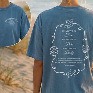 May include: Blue t-shirt with white text and seashell graphics. The front has "Philippians 4:8" and a seashell. The back features a seashell design with the words "Whatever is True, Pure, Lovely".