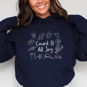 May include: Navy blue hoodie with a front pocket and drawstring. The hoodie features a white graphic with the text "Count It All Joy" surrounded by ocean-themed illustrations, including a sun, seashells, and a starfish.