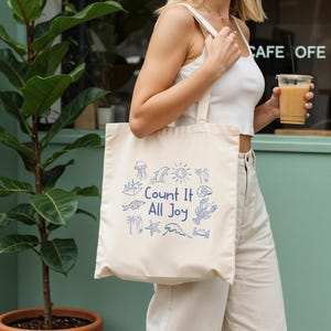 May include: Cream-colored tote bag with blue illustrations of marine life and the text "Count It All Joy." The bag has a shoulder strap and is being held by a person. The background includes a green wall and a potted plant.