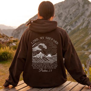 May include: A brown hoodie with the text "LORD, MY SHEPARD" and a graphic of waves and a sun. The text "Psalms 23" is also visible. The hoodie is worn outdoors with a mountain backdrop.