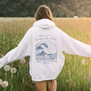 Beautiful woman showcasing the back of a white hoodie. The bible verse, Isaiah 43 2 scripture with illustration of waves in light blue