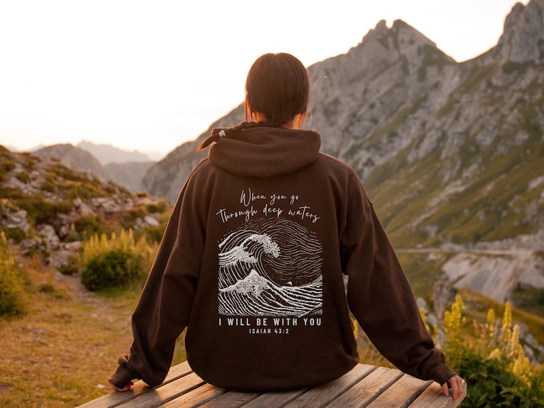 Beautiful woman showcasing the back of a dark chocolate hoodie. The bible verse, Isaiah 43 2 scripture with illustration of waves in white