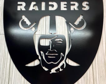 Puede incluir: Letrero negro de metal con forma de escudo de los Raiders. La palabra "RAIDERS" está recortada en la parte superior. El logotipo de los Raiders, una cara con casco y espadas cruzadas, está en el centro. El letrero está montado sobre un fondo claro de vetas de madera.