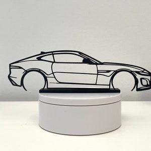 Puede incluir: Una silueta de coche deportivo de metal negro sobre un pedestal blanco. El coche está en vista de perfil y tiene un diseño elegante y moderno.