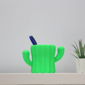 May include: A bright green cactus-shaped desk organizer with two blue pens sticking out of the top. The organizer is sitting on a white surface.