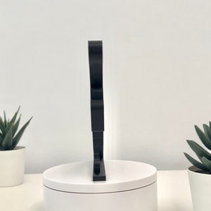 May include: A black, freestanding, three-dimensional letter "S" made of acrylic. The letter is standing on a white circular base.