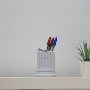 Puede incluir: Un portaplumas de cerámica blanca con forma de columna con tres bolígrafos en su interior. Los bolígrafos son azules, verdes y rojos. El portaplumas está sobre una superficie blanca.