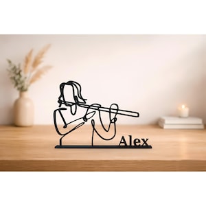 Op de afbeelding: Zwarte metalen sculptuur van een persoon die fluit speelt. De sculptuur is een doorlopend lijnontwerp met de naam "Alex" aan de basis. De sculptuur staat op een houten oppervlak.