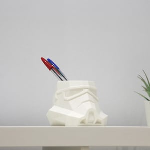 Puede incluir: Porta-bolígrafos de plástico blanco con forma de casco de Stormtrooper de Star Wars. El casco está sobre una superficie blanca con dos bolígrafos que sobresalen de la parte superior.