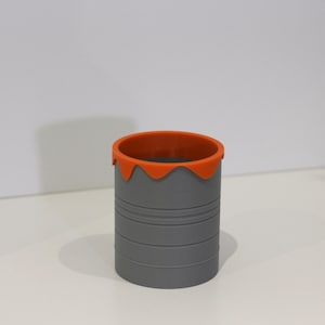 Puede incluir: Un porta lápices de silicona gris con un borde naranja. El borde tiene un efecto de goteo, dando al porta lápices un aspecto de lata de pintura.