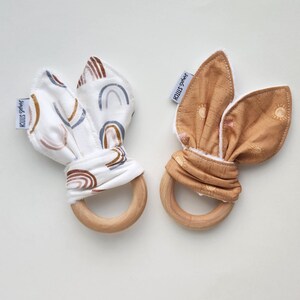May include: Two wooden teethers with fabric coverings. One teether has a white fabric with a rainbow pattern and the text "Hand Stitch". The other teether has a brown fabric with a sun pattern and the text "Hand Stitch".
