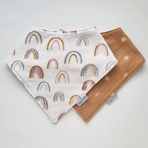 May include: Two baby bibs. One bib is white with a pattern of brown, blue, and tan rainbows. The other bib is tan with a pattern of small suns. Both bibs have a tag that says "Simple Stitch".