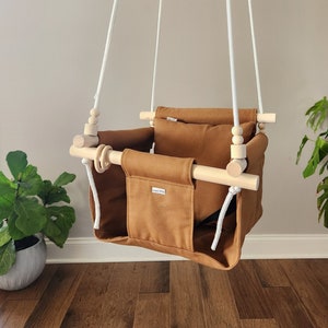 May include: A hanging baby swing made of brown fabric and natural wood. The swing has a pocket on the front and is suspended by white ropes. The swing is designed for indoor use.