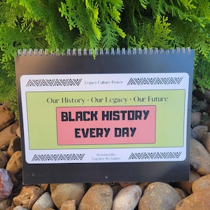 May include: A spiral-bound calendar with the text "BLACK HISTORY EVERY DAY" in a pink box. The calendar has a green and white border with the text "Our History • Our Legacy • Our Future". The calendar is on a bed of rocks.