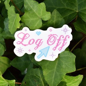 May include: A white sticker with pink text that says "Log Off" with a blue pixelated arrow and white snowflakes. The sticker is on a bed of green leaves.