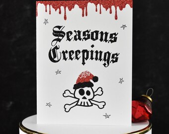 Può includere: Biglietto di auguri bianco con glitter rossi che gocciolano dall'alto, con il testo "Seasons Creepings" in carattere gotico. Un teschio con un cappello da Babbo Natale è sotto il testo, con stelle intorno. Un ornamento rosso è a destra.