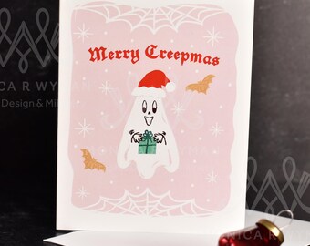 Può includere: Un biglietto di Natale rosa con un fantasma bianco che indossa un cappello da Babbo Natale e tiene un regalo verde. Il biglietto dice "Merry Creepmas" in lettere rosse.