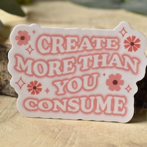 May include: White sticker with pink text that reads "Create more than you consume". The text is in a retro font style. There are pink flowers and sparkles around the text.