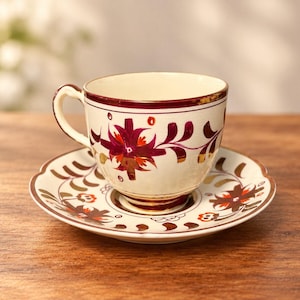 May include: A cream-colored ceramic teacup and matching saucer featuring a hand-painted floral pattern in deep burgundy and orange tones. The cup has a curved handle and a gold-rimmed edge, with matching decorative leaf motifs on the saucer.