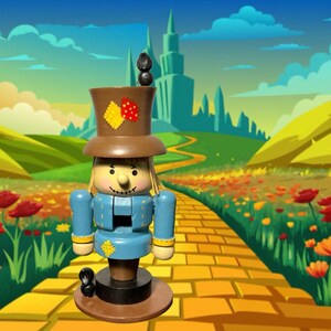 Puede incluir: Un muñeco cascanueces de madera con sombrero de copa marrón y chaqueta azul, de pie sobre una base redonda. El muñeco tiene pelo rubio y una cara sonriente. El fondo presenta un camino de ladrillos amarillos y un paisaje de fantasía.