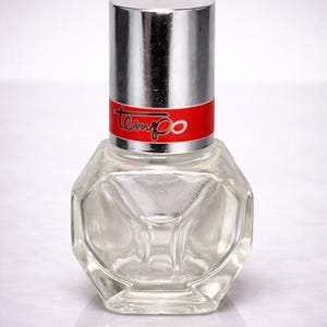 May include: A clear, faceted glass bottle with a silver cap and a red band that reads "tempo" in white. The bottle has a unique geometric shape and reflects light.