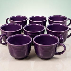 May include: A set of eight deep purple ceramic coffee mugs arranged in three rows. Each mug features a smooth, glossy finish with a series of horizontal concentric rings embossed around the exterior body and a curved handle on the side.