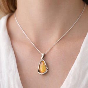 Könnte beinhalten: Eine silberne Halskette mit einem Anhänger, der einen tropfenförmigen Opal zeigt. Der Opal weist orange, gelbe und grüne Farbtöne auf. Der Anhänger ist in eine silberne Lünette gefasst, die Kette wird um den Hals getragen.
