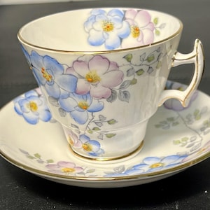 Puede incluir: Una taza de té y un platillo blancos con un patrón floral. La taza y el platillo tienen un borde dorado. Las flores son azules, rosas y verdes.
