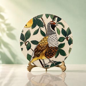 May include: A circular decorative plate featuring a detailed illustration of a quail perched among green leaves and a yellow fruit. The bird has a patterned neck, a golden-brown wing, and a dark head crest. It rests on a gold-toned metal stand.