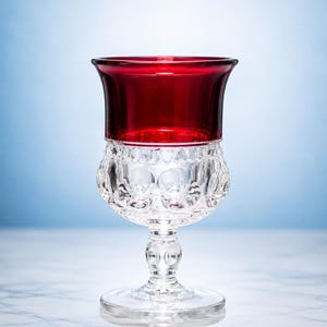 Può includere: Un calice in vetro con una coppa rosso intenso e uno stelo con base trasparenti e incolori. La parte inferiore della coppa presenta un motivo testurizzato a rientranze circolari, mentre il bordo superiore è in vetro rosso liscio.