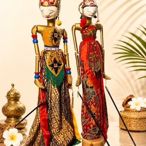 May include: Two Wayang Golek rod puppets with articulated wooden arms and painted faces. The left puppet wears a yellow bodice and patterned skirt, while the right puppet wears a red bodice and floral batik skirt. Both feature ornate headpieces.