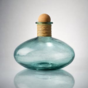 Puede incluir: Una botella de vidrio transparente de color turquesa, con un cuerpo redondeado y un tapón de corcho. El cuello de la botella está envuelto con una cuerda de color natural. La botella se refleja en una superficie blanca.