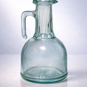 May include: A clear glass jug with a handle, ideal for serving beverages. The jug has a rounded base and a narrow neck with a small spout. The glass has a slight green tint, and the jug is sitting on a white surface.