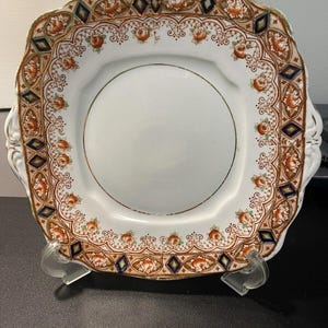 May include: Square white plate with a decorative border featuring orange floral and geometric patterns. The rim has a gold trim. The plate is supported by a clear plastic stand. The plate has a vintage aesthetic.