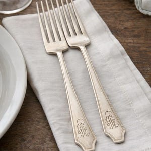 May include: Two silver forks with a monogram design rest on a light gray linen napkin. The forks have long, slender handles and tines. The background includes a wooden table, a white plate, and glass salt and pepper shakers.
