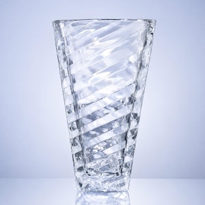 May include: A clear glass vase featuring a distinct diagonal ribbed pattern across its surface. The vase has a wide, open top that tapers down to a narrower, square-shaped base, creating a geometric silhouette with reflective, textured glass.