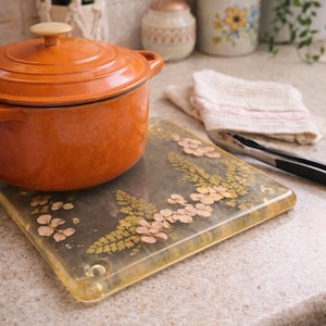 May include: An orange cast iron pot with a lid sits on a decorative floral trivet. The trivet is square and has a clear, yellow border. The background includes kitchen utensils and a white cloth.