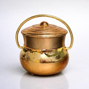 May include: A small, antique-style copper pot with a lid and a handle. The pot has a rounded base and a cylindrical upper section. The lid is domed with concentric rings. The handle is a curved bar. The pot has a golden finish.