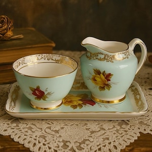 May include: A vintage tea set in light blue, comprising a creamer, sugar bowl, and serving tray. The set is adorned with gold trim and floral patterns. The tray is approximately 20 cm long.