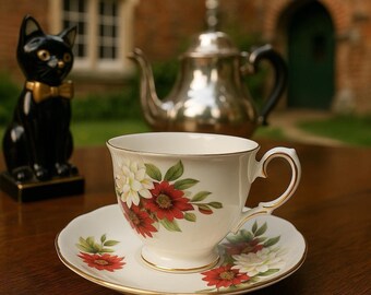 May include: A white teacup and saucer set with a floral design featuring red and white flowers and green leaves. The teacup and saucer have gold trim. A black cat figurine with a gold bow tie is to the left, and a silver teapot is in the background.