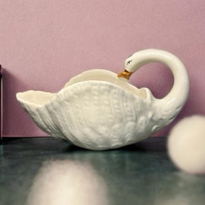May include: A white ceramic swan-shaped serving dish. The swan's neck forms a handle, and the beak is accented with gold. The dish has a textured, woven pattern on the body and sits on a dark surface against a pink background.