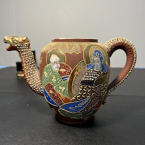 Op de afbeelding: Een bruine theepot van keramiek met een drakenuitloop en -hendel. De theepot is versierd met gouden en zilveren verf en toont een scène met twee mensen in traditionele Japanse kleding.