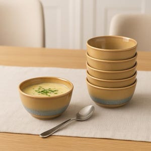 May include: A stack of six tan ceramic bowls with a blue-gray band at the base. One bowl contains soup garnished with green herbs. A silver spoon rests on a white table runner.