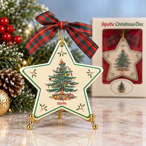 May include: A star-shaped ceramic ornament featuring a Spode Christmas Tree graphic, green trim, and a red and green plaid ribbon. The ornament rests on a gold-toned metal stand, with a matching box labeled Spode Christmas Tree in the background.